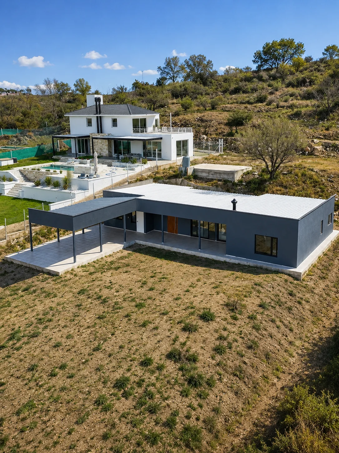 Altos de Carlos Paz — Vista aérea de la casa gris en L con su galería, en un lote amplio; detrás asoma una casa vecina blanca