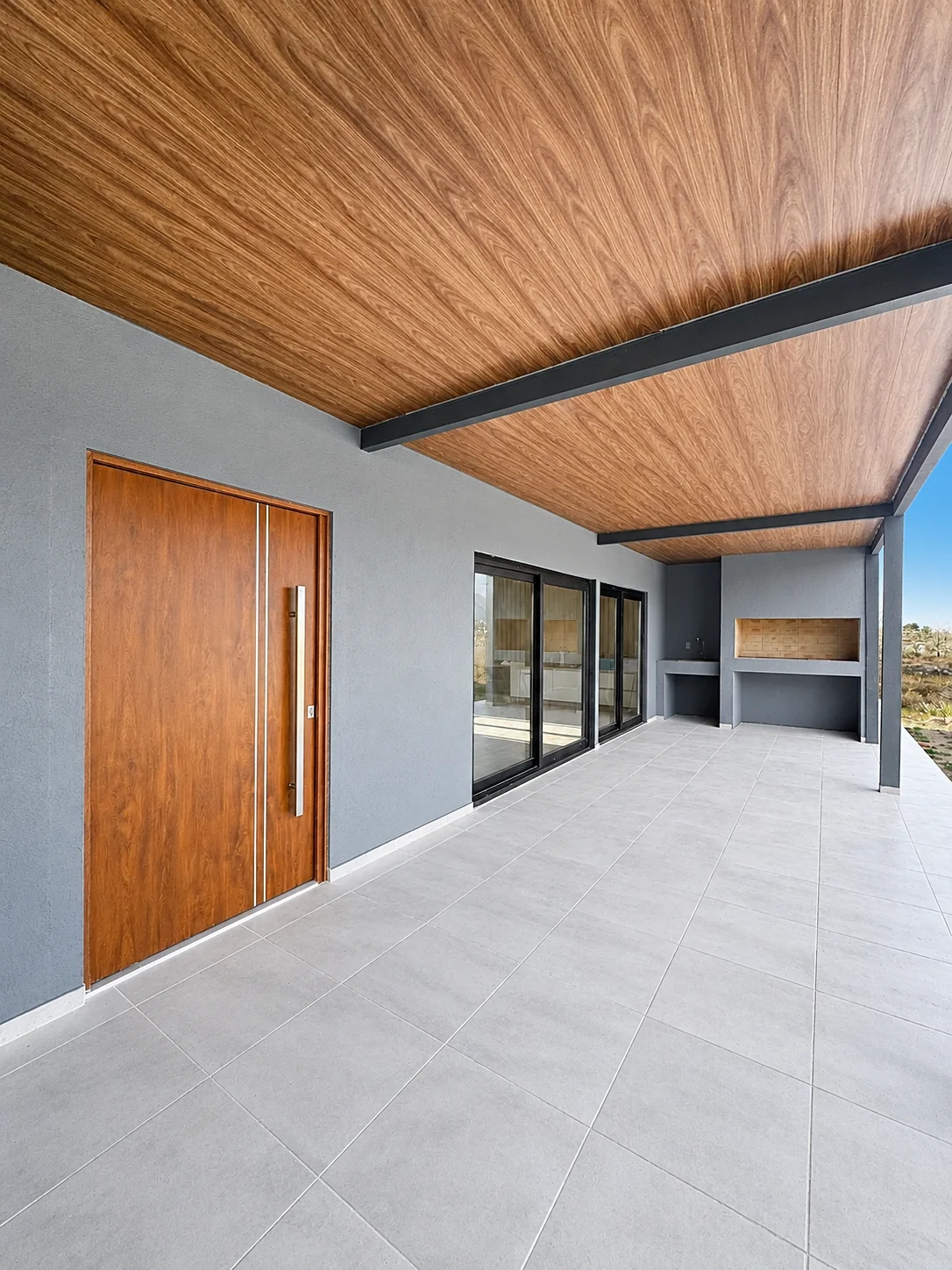 Altos de Carlos Paz — La galería junto a la puerta de madera, con ventanales y la parrilla al fondo