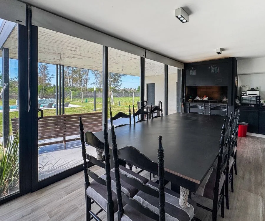 La Melinca Country Golf — Comedor de mesa larga junto a los ventanales; al fondo, la parrilla