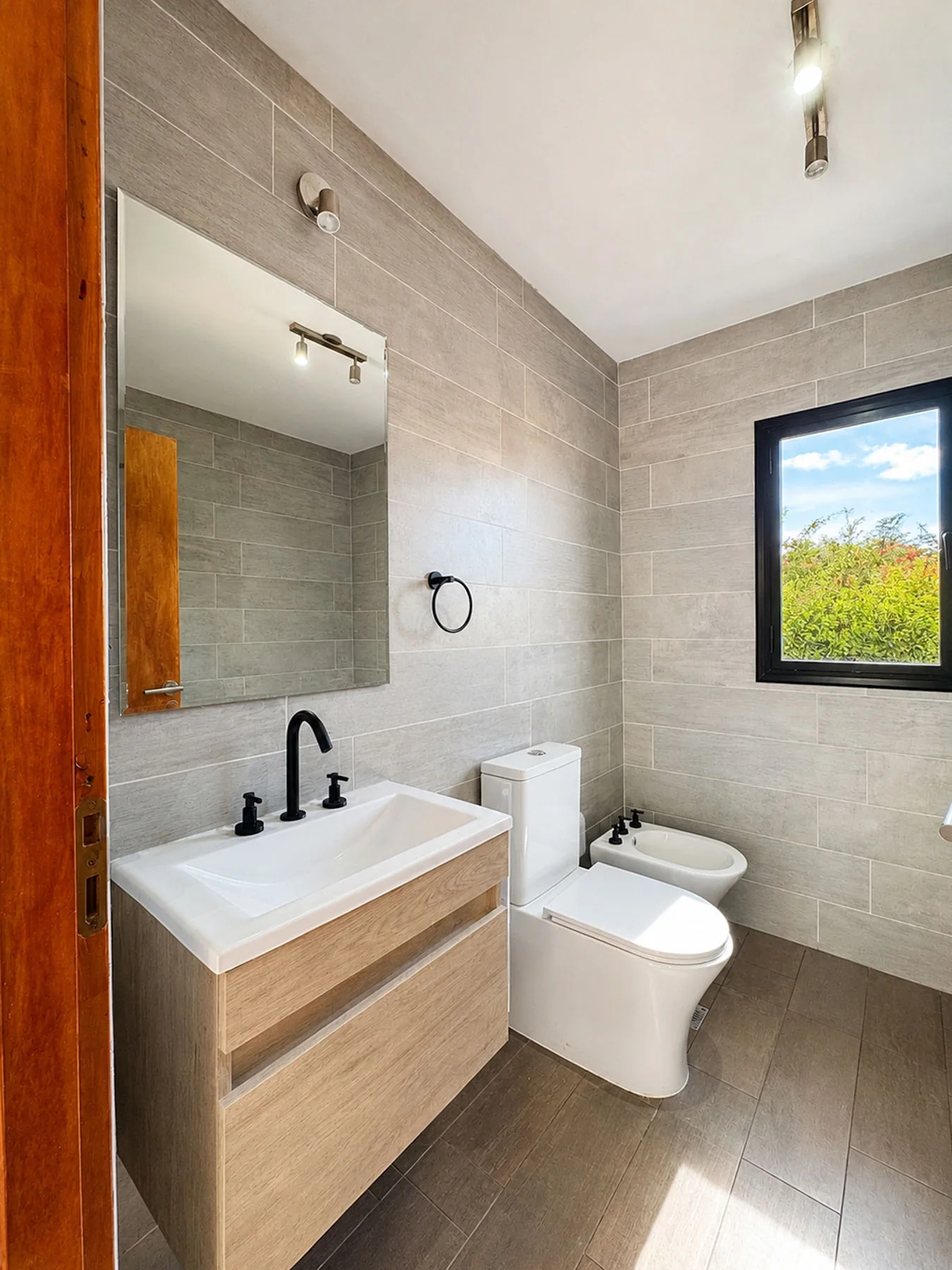 Villa Giardino — Baño revestido en gris, con vanitory, inodoro, bidet y ventana