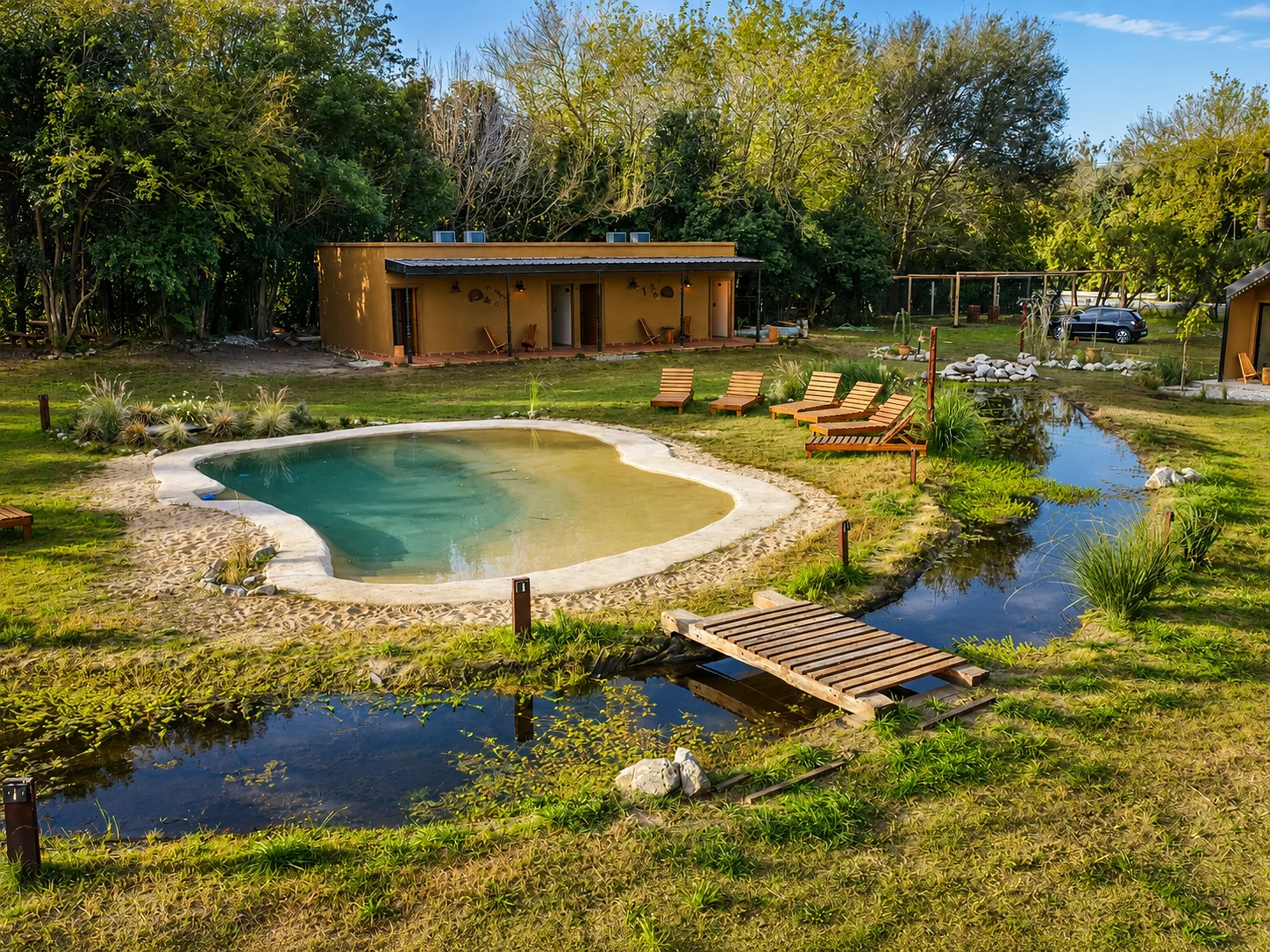 Villa Vadori · Suites Serranas — La pileta, el arroyo con su puente de madera y, al fondo, el edificio largo con galería