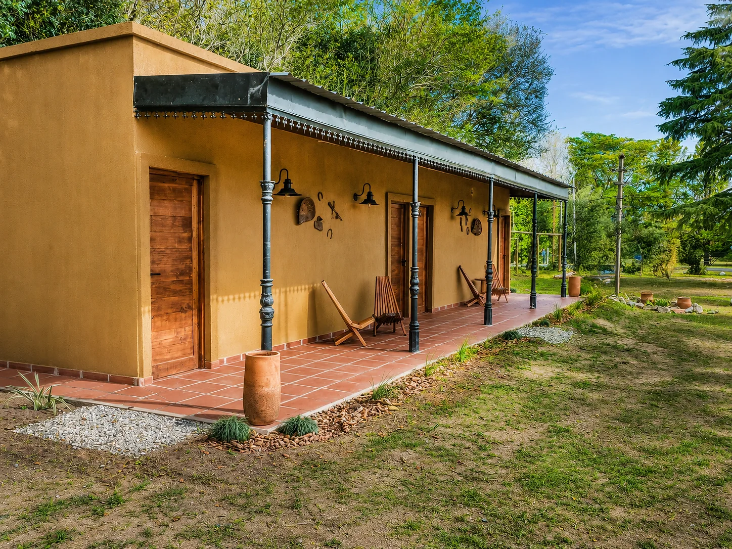 Villa Vadori · Suites Serranas — Edificio largo con galería de columnas negras, piso cerámico rojo y puertas de madera