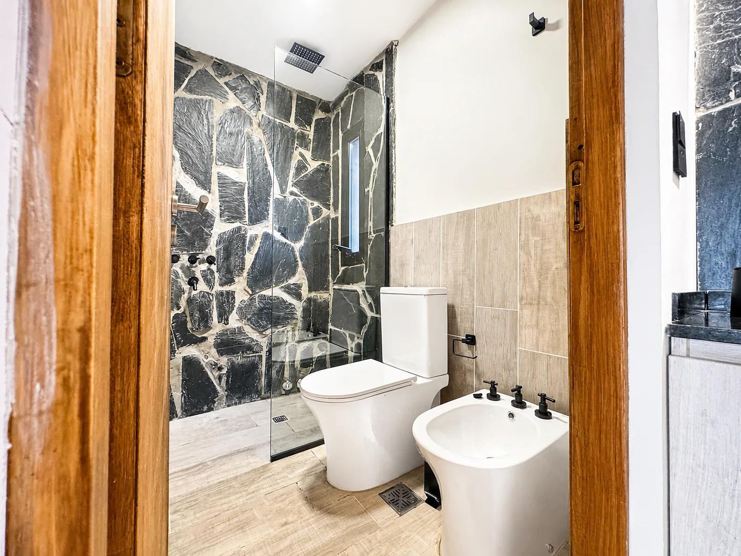 Villa Vadori · Suites Serranas — Baño de la suite: ducha con mampara y pared de piedra, inodoro y bidet